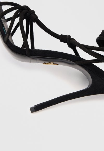 Svart högklackad sandal med korsade mockaband, spetsig tå, slät glänsande sula och en guldaccentdetalj på innersulan.