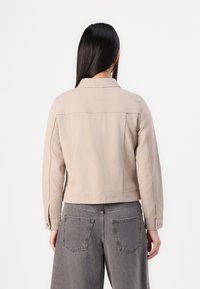 Beez värvi denim jakker, millel on krae, seljaosa talje ja pikad varrukad, kombinatsioonis kõrge vöökohaga hallide laia säärega pükstega.