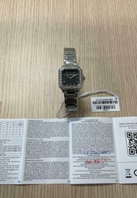 Silberfarbene metallische Uhr mit schwarzem quadratischem Ziffernblatt, Kristallakzenten und einem Gliederarmband, begleitet von einer Garantiekarte und Benutzerinformationen.