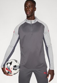 Γκρι και ανοιχτό γκρι αθλητική μπλούζα με φερμουάρ τετάρτου μήκους, σετ με αντίστοιχο παντελόνι. Διαθέτει λογότυπο Puma σε κόκκινο χρώμα· κρατάει μια ασπρόμαυρη μπάλα ποδοσφαίρου.