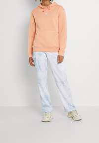 Růžová mikina s kapucí, přední kapsou a stahovací šňůrkou, doplněná světle modrými kalhotami s batikovaným vzorem a neutrálními teniskami.