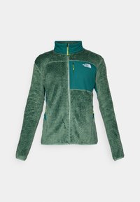 Fleecejakke i mørkegrøn med en textureret overflade, fuld lynlås, høj krave og en lynlåslomme på brystet i kontrasterende teal.