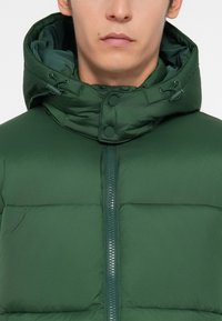 Doudoune verte avec un col montant et une capuche ajustable. Dispose d'une fermeture éclair à l'avant et de boutons-pression, fabriquée en tissu lisse et brillant.
