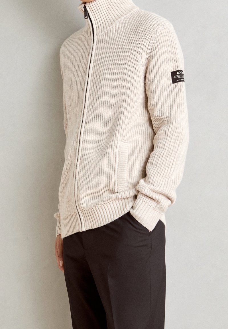 Pull zippé en maille côtelée beige avec col haut et écusson sur la manche gauche, porté avec un pantalon noir, mains partiellement dans les poches.
