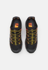 Sapatos atléticos pretos com textura em malha, atacadores amarelos e vermelhos e uma sola robusta. Apresenta um padrão listrado distinto nos atacadores.