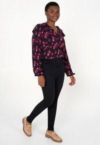 Blouse noire à motifs floraux avec des accents roses, col froncé, et manches longues, assortie à un pantalon noir ajusté et des mocassins beiges avec un détail en métal.