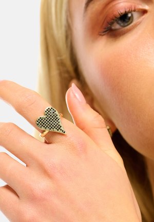 Bague en ton or avec un design en forme de cœur serti de petites pierres noires, portée sur une main à teint clair avec un vernis neutre.