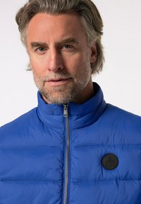 Blå puffjacka med hög krage, vertikal sömnad, dragkedjestängning och en rund svart logotyp på bröstet. Mjukt, isolerat material.