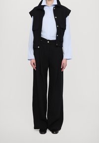 Zwarte wijde broek gecombineerd met een lichtblauw gestreept shirt met lange mouwen en een mouwloos zwart vest met drukknopen.