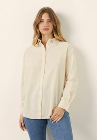 Camisa de manga larga beige claro con cuello abotonado, hecha de una tela texturizada; presenta un corte relajado y puños abotonados.