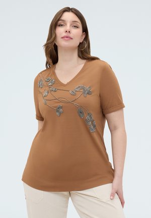 Frau trägt ein braunes V-Ausschnitt-Shirt mit perlenbesetztem Blumenmuster auf der Brust und beige Hose, steht vor einem einfarbigen hellgrauen Hintergrund.