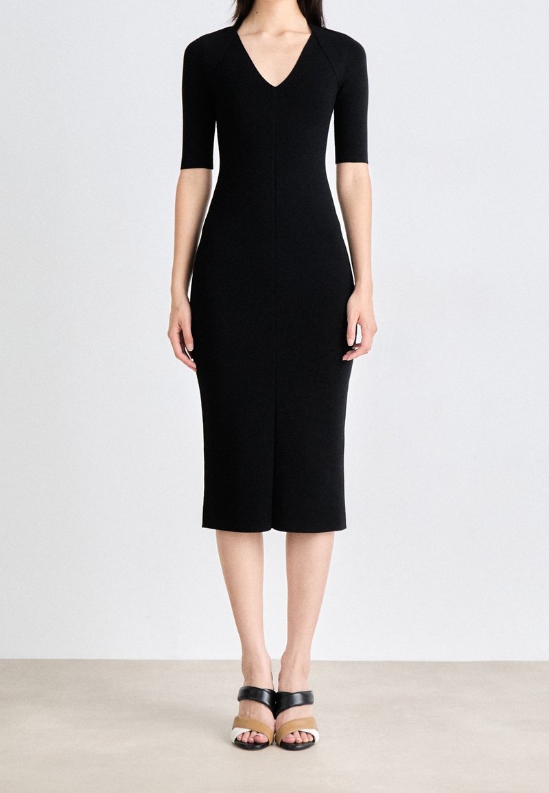 Femme portant une robe midi noire ajustée avec des manches mi-longues jusqu'au coude et un décolleté en V, associée à des sandales à talons bicolores.