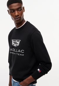 Άνδρας με μαύρο φούτερ με το λογότυπο της ομάδας Cadillac Formula 1 στο στήθος και το λογότυπο σημαίας Tommy Hilfiger στο μανίκι, κοιτάζοντας προς τα κάτω αριστερά.