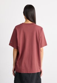 Camiseta de algodón color marrón con mangas cortas y cuello redondo. Presenta un pequeño logotipo de Nike en la manga izquierda. Textura suave.