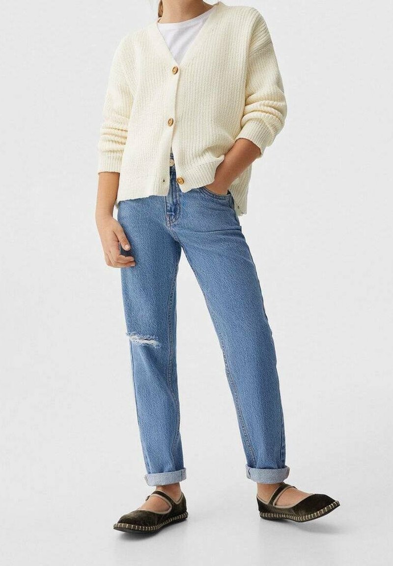 Person som bär en krämfärgad knappad cardigan, vit skjorta, uppvikta slitna blåjeans och mörka slip-on skor, står med ena handen i fickan.