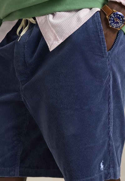 Calções de bombazina azul-marinho com nervuras verticais, bolsos laterais e um pequeno logótipo branco na bainha. Usados com uma camisa verde e colarinho com riscas cor-de-rosa.