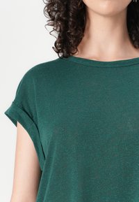 Žalia trumpomis rankovėmis t-shirt maikutė su pasukta rankovės apačia, apvaliu iškirpimu ir laisvu siluetu. Medžiaga atrodo minkšta su švelniu tekstūra.