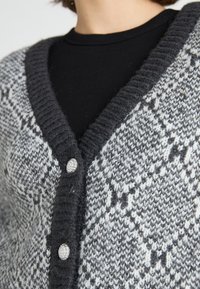 Grijs en wit gemêleerd gebreid vest met een v-hals, voorzien van een dikke ribbelkraag en twee juweelknopen aan de voorkant.