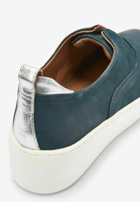 Dunkelgrüner Leder-Schuh mit einem silbernen metallischen Akzent an der Ferse, ausgestattet mit einer weißen Gummisohle und einer glatten Textur.