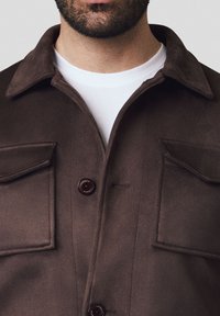 Brązowa zamszowa kurtka z kołnierzem, dwoma przednimi kieszeniami i zapięciem na guzik. Gładka tekstura, dopasowany krój i subtelne detale wykończenia.