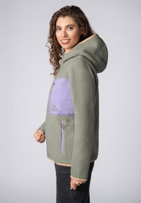Fleece jack in gedempte groene kleur met een lichtpaarse borstzak, verstelbare capuchon en ritssluitingen. Gestructureerde stof met een zachte afwerking.