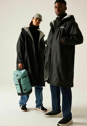 Dos personas llevan abrigos negros largos con capucha y forro de piel, vaqueros azules y zapatillas negras, una de ellas sostiene una mochila azul claro.