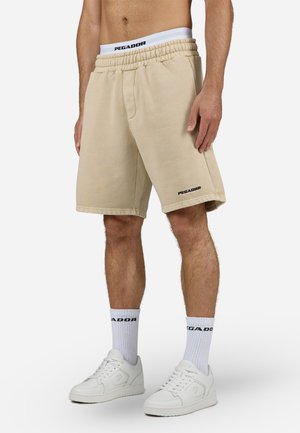 Mężczyzna ubrany w beżowe szorty Pegador, białe skarpetki z logo Pegador oraz białe trampki, stojący na neutralnym tle.