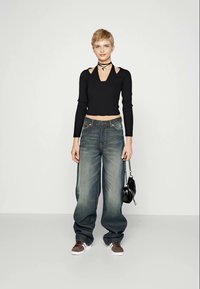 Jeune personne avec des cheveux blonds courts portant un haut découpé noir, un jean évasé, des baskets marron, un choker noir et tenant un petit sac à main noir.