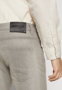 Gray denim jeans with a smooth texture, featuring a leather label with "INDICODE EST. 1993" on the back, and a single back pocket.