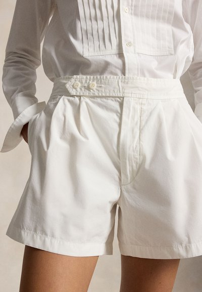 Λευκά σορτς με πιέτες, με ψηλή μέση και κουμπωτή πλαϊνή ταινία, συνδυασμένα με λευκό πουκάμισο μακρυμάνικο, φορεμένο μέσα στη μέση, με τα χέρια στις τσέπες.