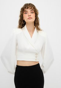 Blusa blanca recortada con mangas largas y abullonadas y escote en V. Presenta tres botones dorados en el frente y una tela suave y estructurada.
