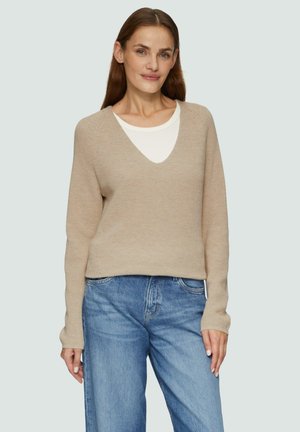 Beżowy sweter dziergany z dekoltem w serek, długimi rękawami i luźnym krojem, nałożony na biały t-shirt, w zestawieniu z jasnoniebieskimi dżinsami.