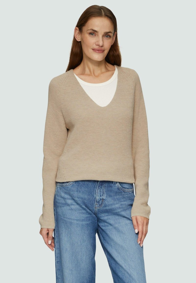 Beige gestrickter Pullover mit V-Ausschnitt, langen Ärmeln und lockerer Passform, über einem weißen T-Shirt getragen, kombiniert mit hellblauen Jeans.