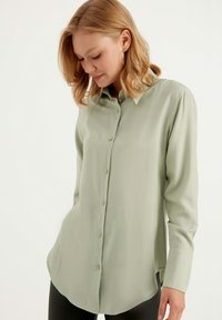 Camicia verde pallido con bottoni, realizzata in tessuto leggero, con colletto classico, maniche lunghe e orlo curvo. Texture liscia e vestibilità sartoriale.