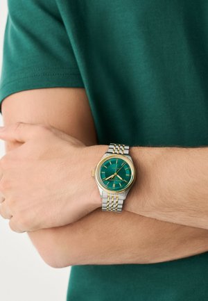 Man draagt een zilveren en gouden polshorloge met een groen wijzerplaat en gouden markeringen, armen gekruist over een groen shirt met korte mouwen.