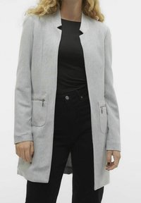 Světle šedý kabát ve herringbone vzoru s širokým límcem, dvěma předními kapsami na zip, dlouhými rukávy a rovného střihu. Nositý přes černý top.