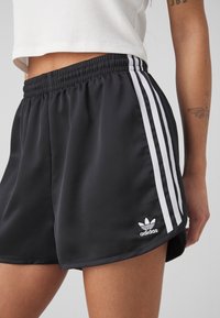 Černé sportovní šortky vyrobené z hladké tkaniny, s bílými bočními proužky, elastickým pasem a malým bílým logem Adidas.