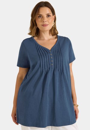 Mujer que lleva una blusa azul marino holgada de manga corta con pliegues y botones en la parte delantera, combinada con pantalones blancos, pendientes dorados y pulseras.
