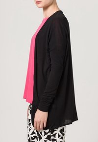 Czarny kardigan na długi rękaw o luźnym kroju i z otwartym przodem, noszony na jasnoróżowym topie oraz wzorzystych czarno-białych spodniach.