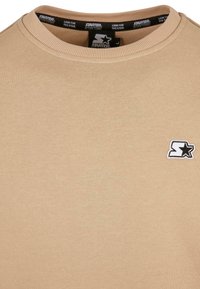 Sudadera beige de cuello redondo con un pequeño logo en blanco y negro de Starter en el lado izquierdo del pecho y cinta de cuello interior de marca.