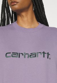 Sweatshirt violet avec un col rond et des poignets côtelés, orné d'un logo "carhartt" brodé en vert foncé sur la poitrine. Texture de tissu douce.