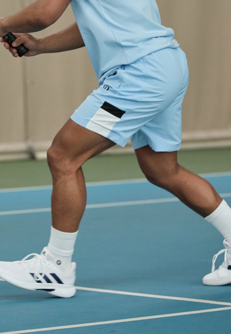 Joueur de tennis en short et chemise bleu clair effectue une fente sur un court intérieur, tenant une raquette, avec des baskets blanches et des chaussettes visibles.