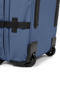 Maleta azul con ruedas, superficie de tela texturizada, acentos de plástico negro, un asa superior y ruedas que giran suavemente.