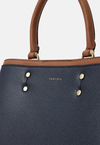 Sac à main en cuir bleu marine avec des accents marron, comportant deux clous métalliques dorés et un logo, des poignées arrondies et une surface texturée.