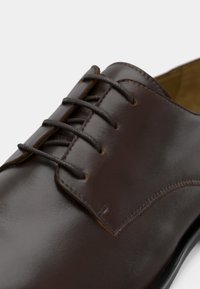 Καφέ δερμάτινο παπούτσι για επίσημες εμφανίσεις με λεία υφή, στρογγυλή μύτη και έξι τρύπες για κορδόνια του ίδιου χρώματος. Διακριτικές ραφές στο επάνω μέρος.