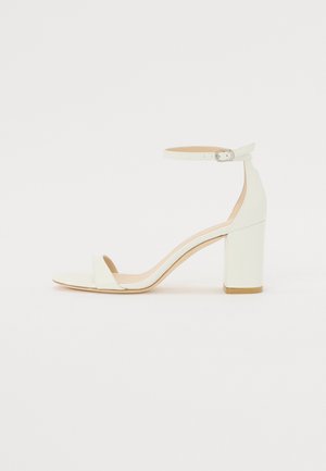 Sandal de bloco em pele branca, apresentando uma correia no tornozelo com fivela e um design minimalista. Com biqueira aberta e texturas suaves ao longo de toda a peça.