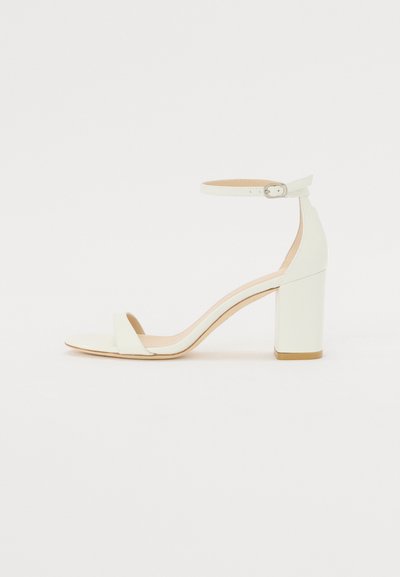 Sandale à talon bloc en cuir blanc avec une bride de cheville à boucle et un design minimaliste. Bout ouvert et textures lisses sur l'ensemble.