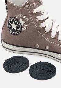 Brown canvas high-top sneaker with white laces, featuring a black and white star logo patch. Two black shoelaces are placed nearby.