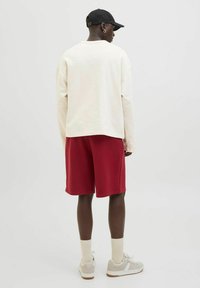 Camisa de manga larga color crema claro, pantalones cortos rojos, gorra negra y zapatillas blancas con detalles en gris. Estilo casual, corte relajado con costuras visibles.