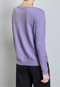 Person wearing a lavender long-sleeve sweater and black pants, standing with their back to the camera against a plain background.